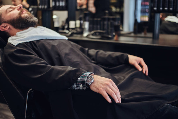 Красивий бородатий чоловік чекає на перукаря, який сидить на перукарському стільці
. - Фото, зображення