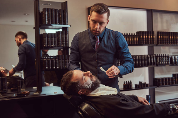 Um belo hipster barbudo sentado numa poltrona numa barbearia enquanto o cabeleireiro rapa a barba com uma perigosa navalha.
. - Foto, Imagem