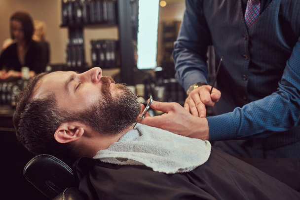Maschio barbuto seduto in poltrona in un negozio di barbiere mentre il parrucchiere modella la barba con le forbici e pettine al barbiere
. - Foto, immagini