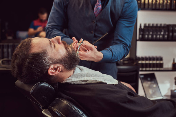 Varón barbudo sentado en un sillón en una peluquería mientras modela la barba con tijeras y peina en la peluquería
. - Foto, Imagen