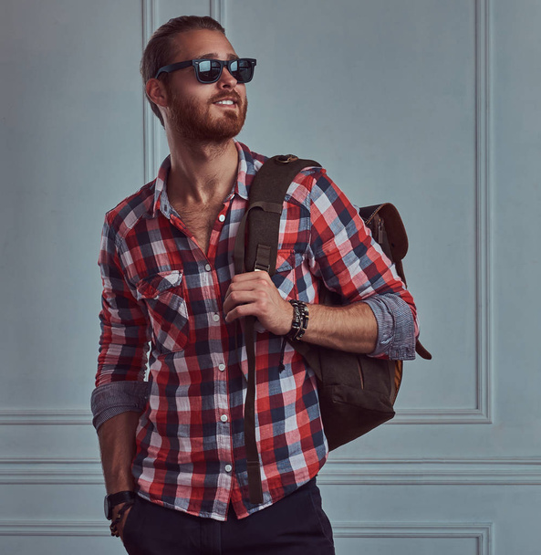 Przystojny uśmiechający się stylowe rude podróżnik w flanelową koszulę i okulary przeciwsłoneczne z plecakiem, pozowanie Studio pod ścianą, biały. - Zdjęcie, obraz