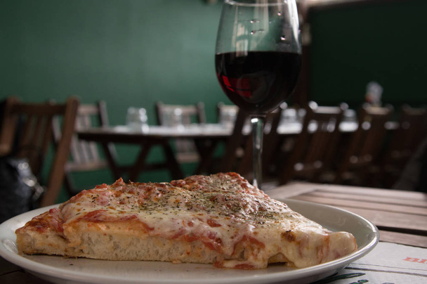 Кусочек пиццы на тарелке с красным бокалом вина в интерьере кафе
 - Фото, изображение