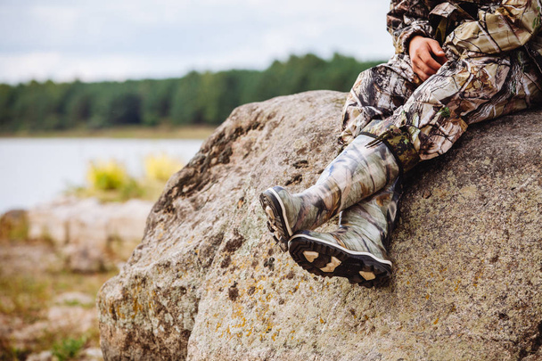 a man in rubber boots and in military uniform sits on a rock near the river in summer - Foto, Imagem