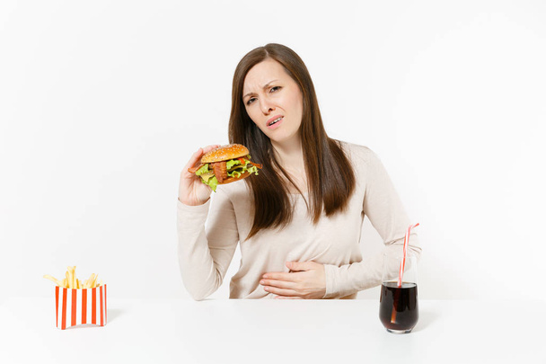 Illness woman put hand on pain abdomen, stomach-ache at table with burger, french fries, cola in glass bottle isolated on white background. Proper nutrition or American classic fast food. Copy space. - Foto, Imagem