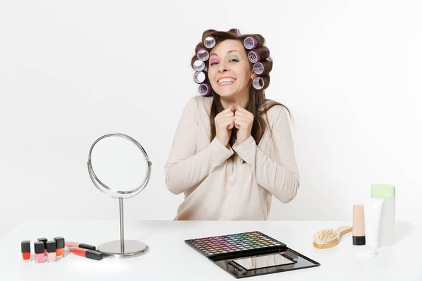 Fun young woman with curlers on hair sitting at table applying makeup with set facial decorative cosmetics isolated on white background. Beauty female fashion lifestyle concept. Area with copy space. - Fotografie, Obrázek