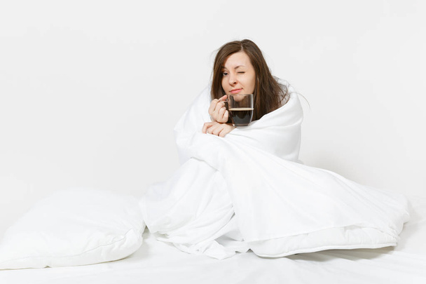 Young tired brunette woman sitting in bed with cup of coffee, white sheet, pillow, wrapping in blanket on white background. Female spending time in room. Rest, relax, good mood concept. Copy space. - Foto, afbeelding