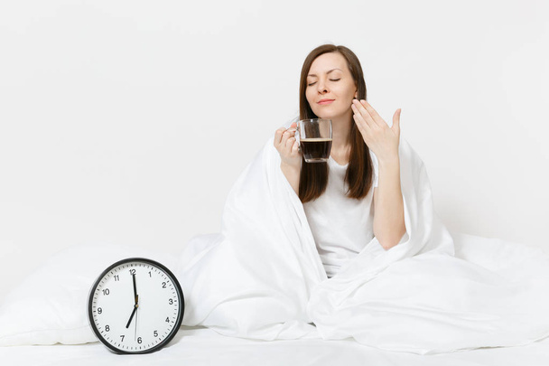 Femme heureuse assise au lit avec horloge ronde, drap blanc, oreiller, enveloppant dans une couverture sur fond blanc. Beauté femelle se réveiller tôt le matin, bénéficie d'un arôme de café. Reste relax, concept de bonne humeur
 - Photo, image