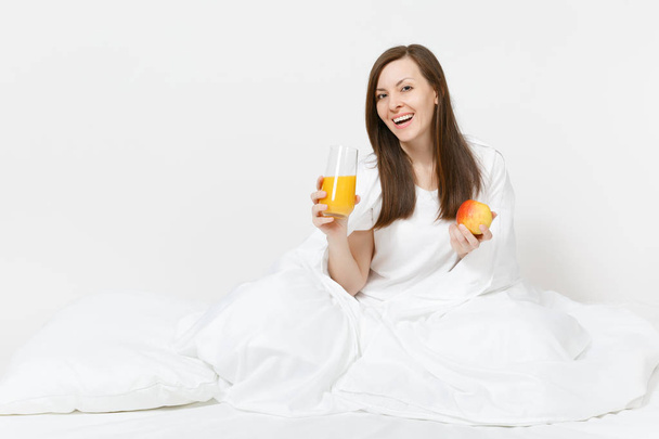 Mujer morena joven se sienta en la cama con sábana blanca, almohada, envoltura en manta aislada sobre fondo blanco. Belleza femenina desayunar con manzana, zumo de naranja fresco. Descanse, relájese, concepto de buen humor
 - Foto, Imagen