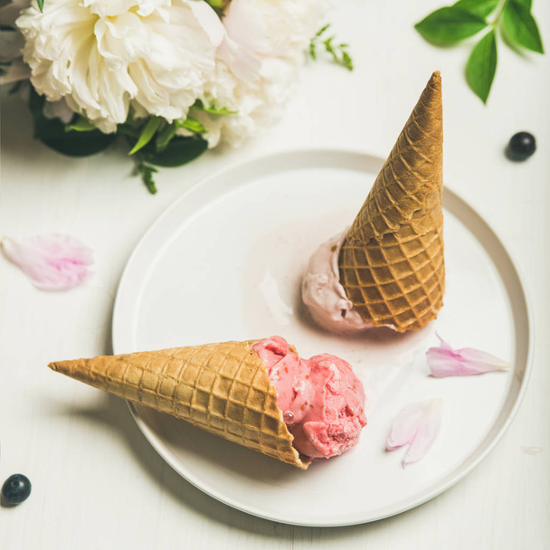 Pink strawberry and coconut ice cream scoops, sweet cones and peony flowers bouquet on plate over white background - Foto, Imagem