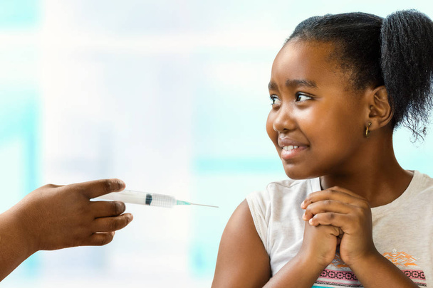 Close up portrait of anxious little african girl scared of syringe with needle.Kid looking aside at doctor with nervous facial expression. Hand holding syringe near upper arm. - Foto, Bild