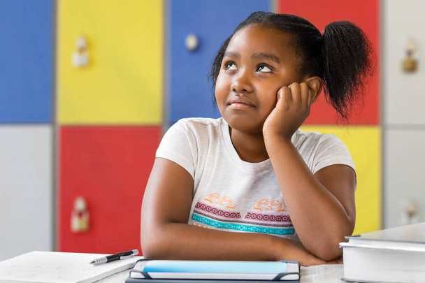 Close up portrait of little african female student sitting at desk in classroom. Tedious kid with bored wondering facial expression staring and looking up. - 写真・画像
