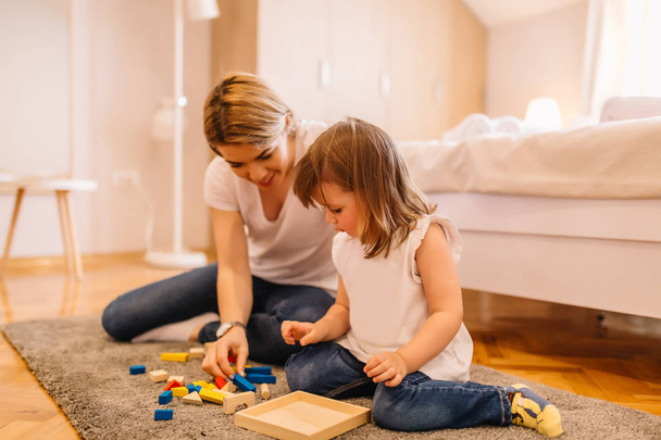 Cute little girl is solving wooden puzzles with her mother on the floor in bedroom - Photo, Image