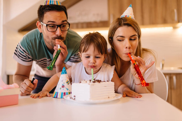 niedliches kleines Mädchen feiert kleine Geburtstagsparty mit ihren Eltern und bläst Kerze auf Geburtstagstorte - Foto, Bild