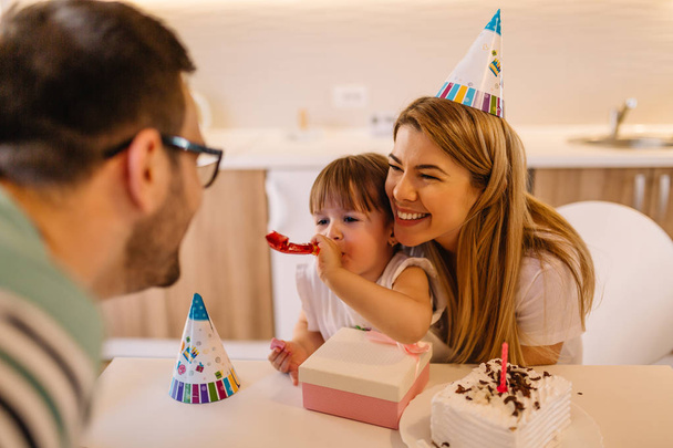 Cute little girl is with her parents in their home and she is blowing a party horn because it is her birthday - Photo, Image