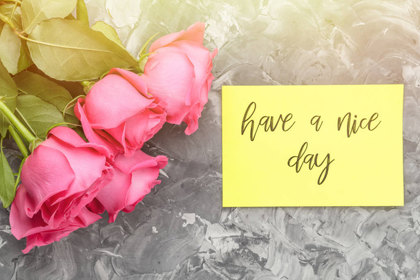 Różowe róże i żółtą kartkę z napisem, milego dnia, szare tło betonu w słońcu. Pojęcie dobry nastrój - Zdjęcie, obraz