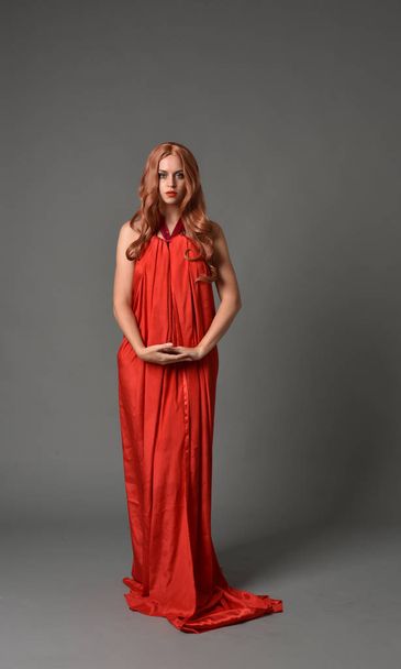 Ολόσωμο Πορτρέτο γυναίκας που φορώντας ένα κόκκινο μεταξωτό φόρεμα, στέκεται πόζα σε φόντο γκρι στούντιο. - Φωτογραφία, εικόνα