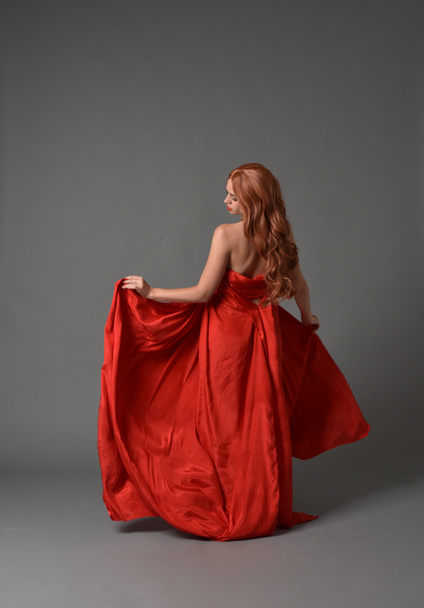 портрет повної довжини жінки в червоній шовковій сукні, стоячи позу на сірому студійному фоні
. - Фото, зображення