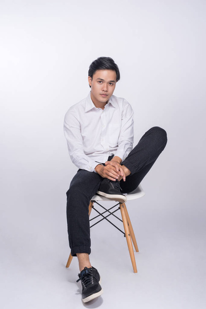 Smart casual asian man seated on chair, posing while looking at camera in studio background - Фото, изображение