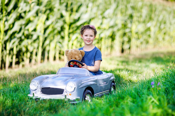 Pequena menina criança pré-escolar dirigindo carro de brinquedo grande e se divertindo com brincar com grande bea brinquedo de pelúcia
 - Foto, Imagem