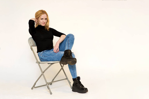 Portrait of a beautiful blonde girl in jeans sitting on a chair on a white background - Φωτογραφία, εικόνα