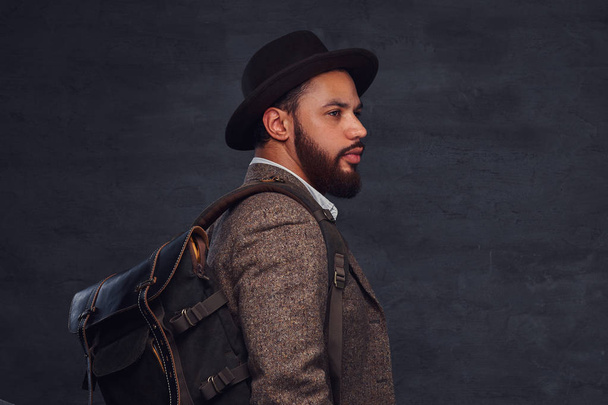 Yakışıklı Afro-Amerikan gezgin bir kahverengi ceket ve şapkayla sırt çantası, bir stüdyoda duruyor. Karanlık bir arka plan üzerinde izole. - Fotoğraf, Görsel