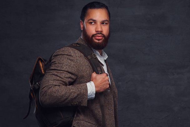 A handsome Afro-American traveler in a brown jacket with the backpack, stands in a studio. Isolated on a dark background. - Photo, Image