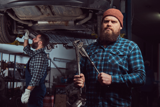 Дві бородаті жорстокі механіки ремонтують машину на ліфті в гаражі
. - Фото, зображення