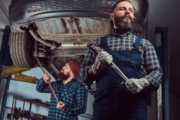 zwei bärtige brutale Mechaniker reparieren ein Auto auf einem Aufzug in der Garage. - Foto, Bild