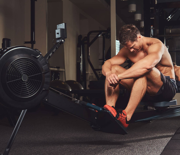 Um atleta sem camisa muscular descansando após um treino duro enquanto se senta na máquina de remo no ginásio
. - Foto, Imagem