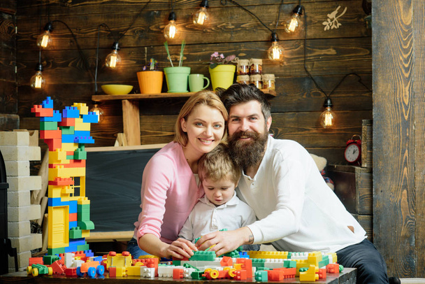 Kid with parents play with plastic blocks, build construction. Father, mother and cute son play with constructor bricks. Family leisure concept. Parents hugs, watching son playing, enjoy parenthood. - 写真・画像