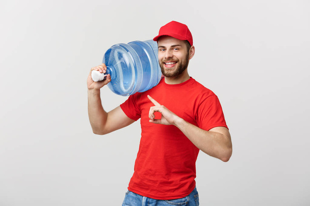 Portrait sourire bouteille d'eau livraison courrier en t-shirt rouge et bouchon portant réservoir de boisson fraîche et doigt pointeur isolé sur fond blanc
. - Photo, image