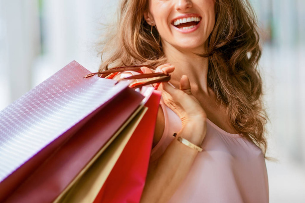 A brown-haired woman holding three shopping bags - brown, gold, and red - over her right shoulder looks back at someone who is making her laugh. A good shopping spree makes any woman happy. She is content, exudes elegance and effortless style. She lo - Photo, Image