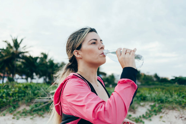 Γυμναστήριο δρομέας γυναίκα πόσιμο νερό από ένα μπουκάλι σπορ. Αθλητής κορίτσι, λαμβάνοντας ένα διάλειμμα κατά τη διάρκεια εκτέλεση στην ενυδάτωση κατά τη διάρκεια άσκησης καυτό καλοκαίρι στην παραλία. Δραστήριου τρόπου ζωής. - Φωτογραφία, εικόνα
