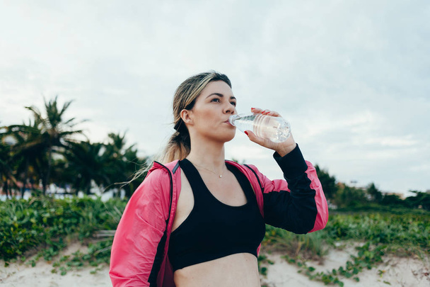 フィットネス ランナー女性飲料水のスポーツ ボトル。中に休憩を取ってアスリート少女をビーチで夏の暑い練習中にメタンハイド レートを実行します。健康的なアクティブなライフ スタイル. - 写真・画像