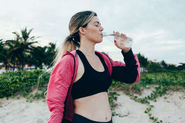 Фитнес-бегунья пьет воду из спортивной бутылки. Спортсмен девушка берет перерыв во время пробежки, чтобы увлажнить во время жарких летних упражнений на пляже. Здоровый активный образ жизни
. - Фото, изображение