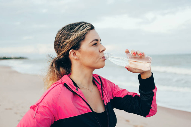 Fitness runner woman drinking water of a sport bottle. Athlete girl taking a break during run to hydrate during hot summer exercise on beach. Healthy active lifestyle. - Zdjęcie, obraz