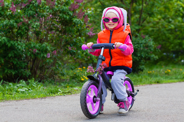 Aktiivinen vaalea lapsi ajaa ajaa polkupyörällä kaupungin katu kujalla. Funny rento vaatteet liivi takki, aurinkolasi ja huppu pupu korvat. Tasapaino pyörä käsite
. - Valokuva, kuva