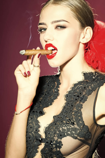 Portrait of one attractive woman in black lace retro clothing and red bow in head with bright lips holding cigar in mouth and smoking looking forward in studio on purple background, vertical picture - Foto, afbeelding