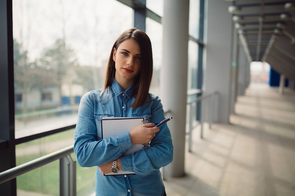 Beautiful brunette woman holding notebook and pencil while standing in corridor - Φωτογραφία, εικόνα