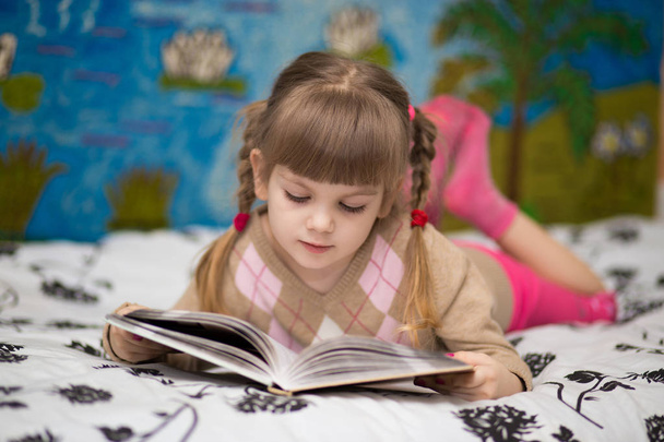 Μικρό χαρούμενο κορίτσι διαβάσει το βιβλίο στο κρεβάτι. Έννοια της γνώσης τα παιδιά - Φωτογραφία, εικόνα
