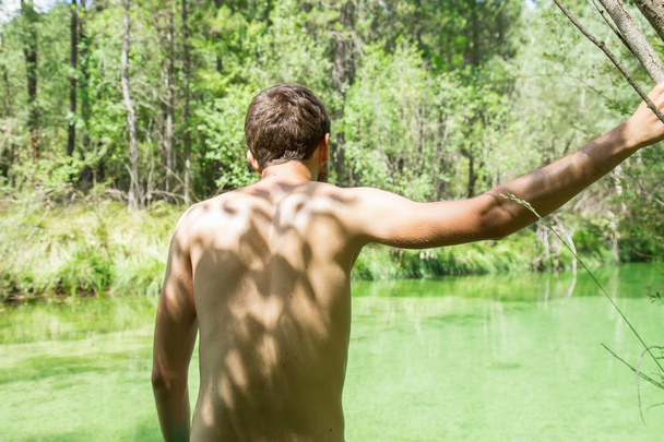 Young man looking at the greenish river a sunny day seen from his back. - Photo, Image