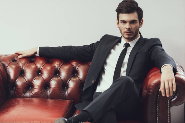 Confident young businessman in suit sitting in red leather armchair isolated. Business style. - Fotografie, Obrázek