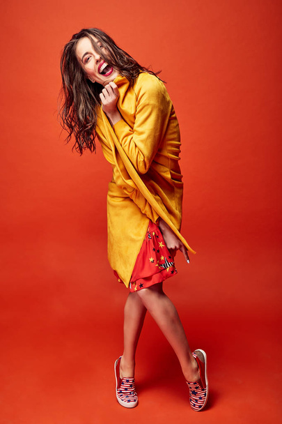 Amazing emotional woman with dark wavy hair, dressed in red star print dress and yellow coat, posing in studio with dark orange background - Photo, image