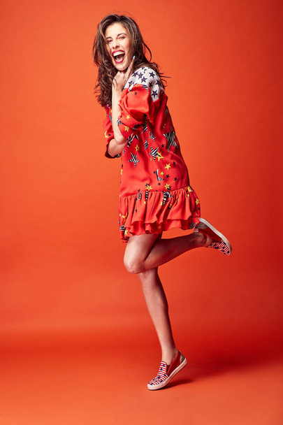 Amazing cheerful woman with dark wavy hair, dressed in red star print dress, posing in studio with dark orange background - Foto, Imagem