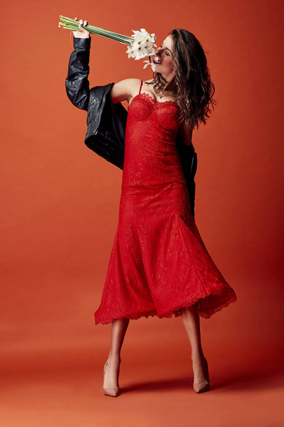 Cheerful brunette woman dressed in sexy red lace dress, black leather jacket and beige heels posing with bunch of daffodils in studio with orange background  - Foto, imagen