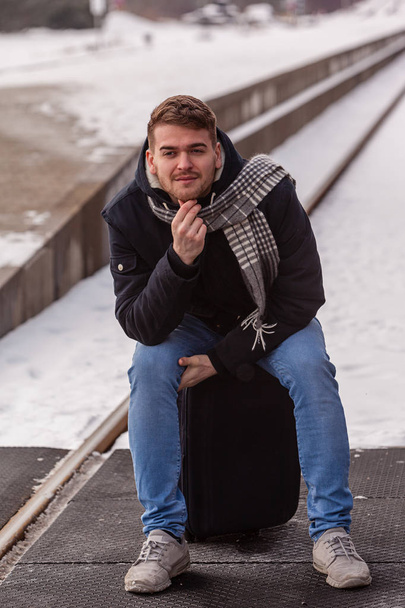 若い男が彼の列車を待っている冬に座って彼のスーツケースで. - 写真・画像
