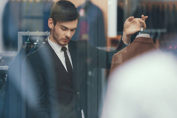 Succesvolle jonge zakenman kijkt jas in business mens kledingwinkel. - Foto, afbeelding