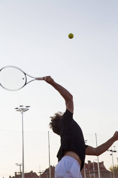 Vista trasera del deportista con raqueta de tenis golpeando la pelota en la luz trasera en la cancha
. - Foto, Imagen