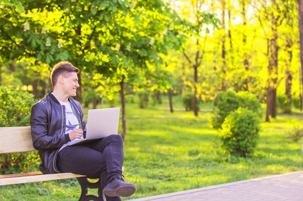A young handsome man is sitting and working in the park with a laptop. The guy freelancer works outside and smile - Foto, Bild