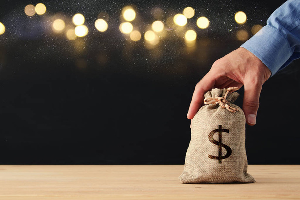 Male hand holding a sack of money over wooden desk and glitter black background. - Photo, image
